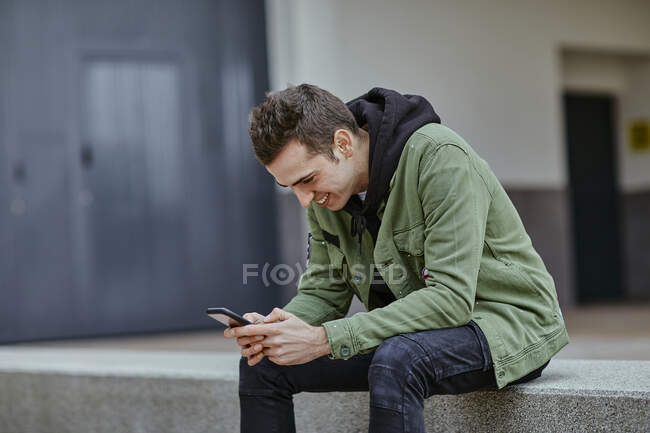 Sonriente hombre guapo usando el teléfono inteligente mientras está sentado en la pared de contención en el café de la acera - foto de stock