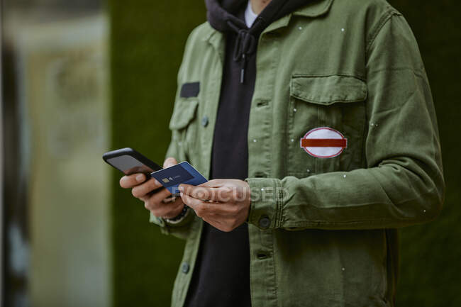 Hombre con chaqueta sosteniendo el teléfono móvil mientras hace compras en línea a través de tarjeta de crédito - foto de stock
