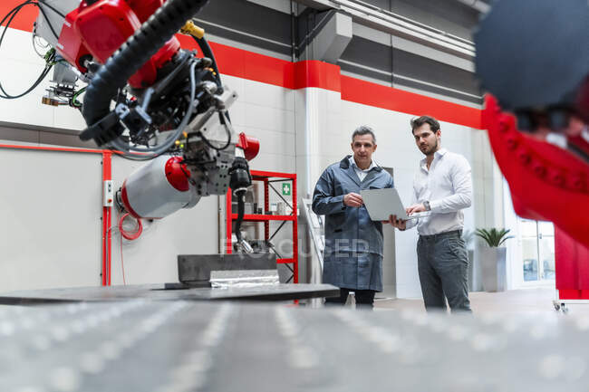 Ingegneri maschi con computer portatile guardando la robotica mentre in piedi in fabbrica — Foto stock