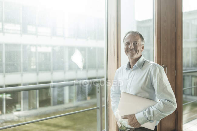 Uomo d'affari sorridente che tiene il computer portatile mentre si trova vicino alla finestra in ufficio — Foto stock