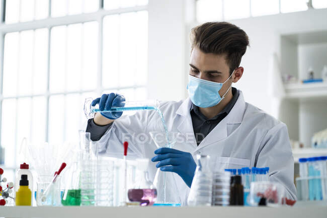 Hombre científico vertiendo líquido en frasco en el laboratorio durante la pandemia - foto de stock