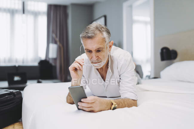Homme utilisant un téléphone intelligent tout couché sur le lit dans la chambre d'hôtel — Photo de stock