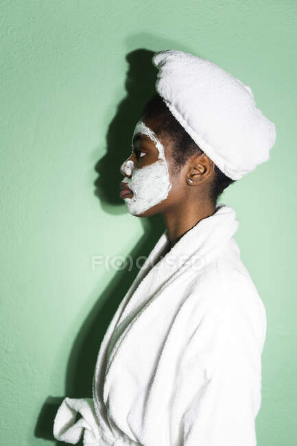Jovem mulher em roupão de banho com máscara facial olhando para longe, enquanto em pé contra o fundo verde — Fotografia de Stock