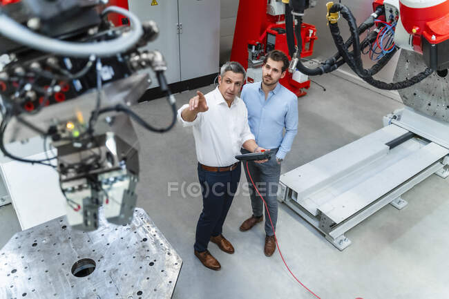 Зрелый мужчина-инженер с управлением, указывающий на робототехнику, стоя рядом с молодым человеком в промышленности — стоковое фото