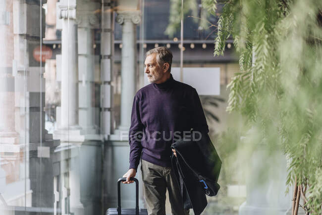 Man with jacket and luggage looking away while walking by glass wall — Stock Photo