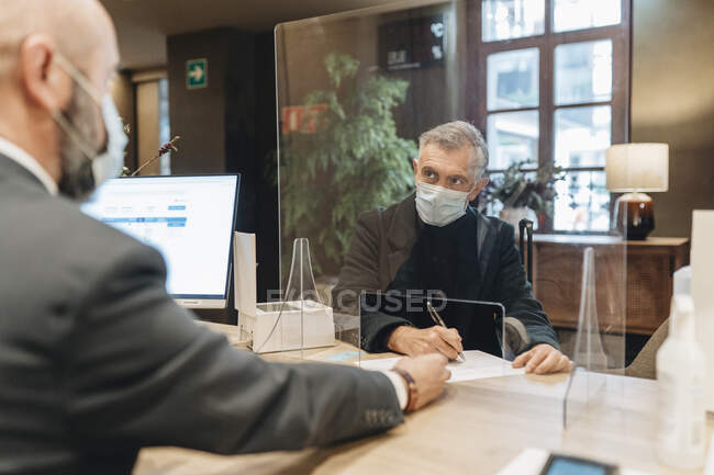 Hombre con máscara protectora llenando formulario en recepción del hotel - foto de stock