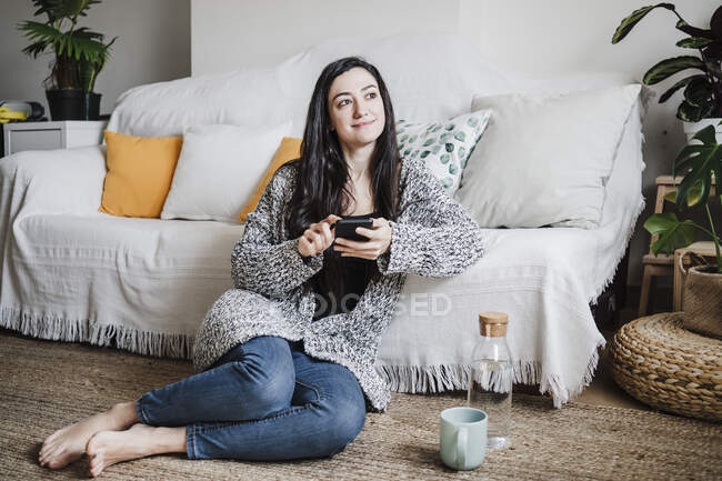 Donna sorridente che distoglie lo sguardo mentre usa lo smartphone contro il divano in soggiorno — Foto stock