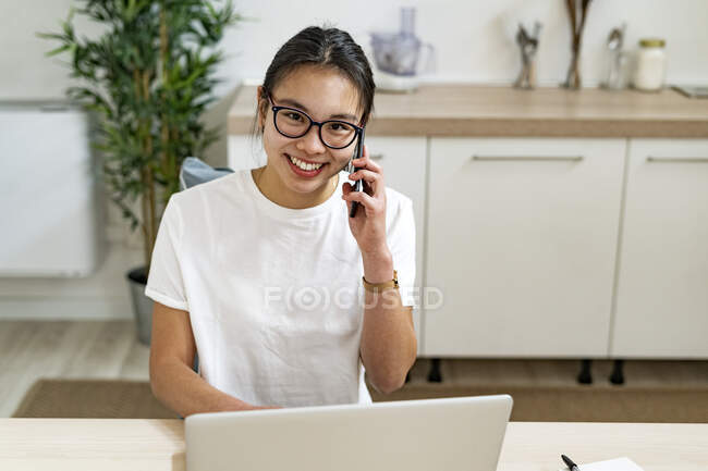 Mujer hablando en el teléfono móvil mientras está sentado con el ordenador portátil en casa - foto de stock