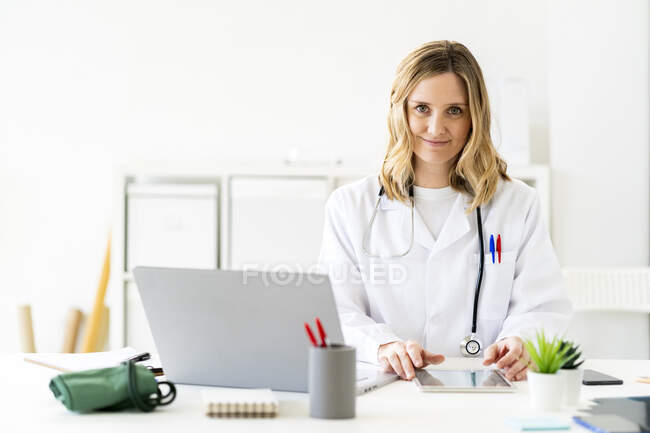 Mujer rubia profesional médico sentado con la tableta y el ordenador portátil en el escritorio - foto de stock