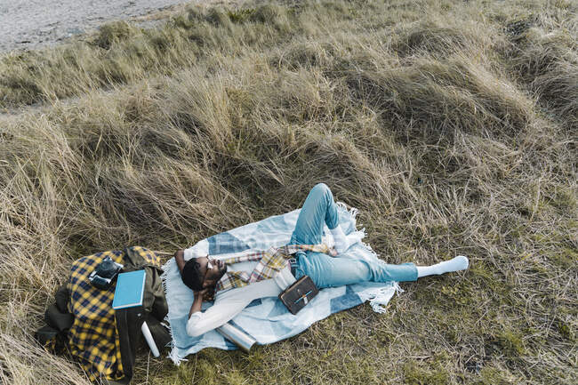 Homem com as mãos atrás da cabeça relaxando em cobertor em meio a planta seca na praia — Fotografia de Stock