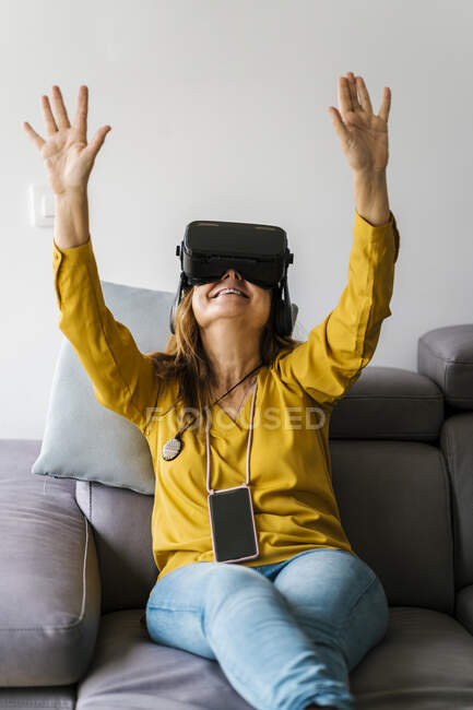 Зрелая женщина жестикулирует в наушниках виртуальной реальности, сидя дома на диване — стоковое фото