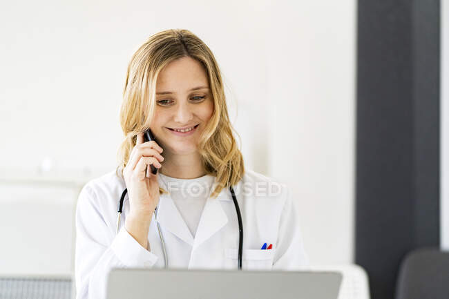 Sonriente doctora rubia hablando por teléfono inteligente en clínica médica - foto de stock