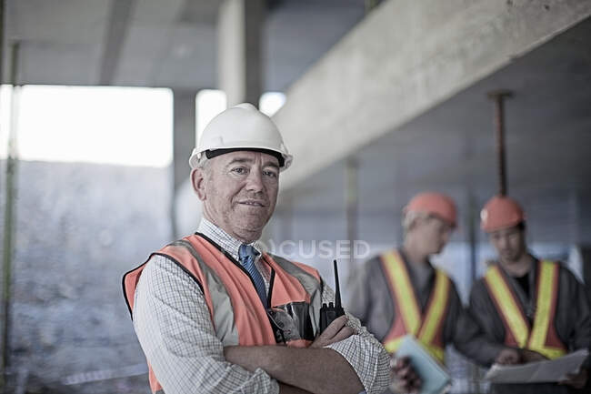 Entrepreneur masculin confiant avec les bras croisés sur le site avec des travailleurs de la construction vus en arrière-plan — Photo de stock