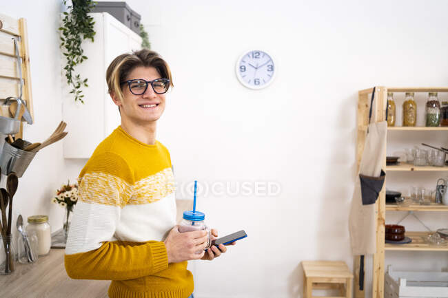 Homme souriant portant des lunettes tenant le téléphone et le pot de smoothie dans la cuisine — Photo de stock