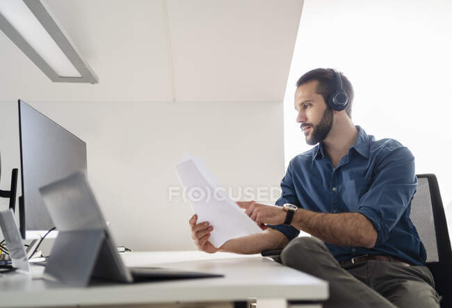Profesional masculino con audífonos comprobando papel mientras usa el ordenador en la oficina - foto de stock