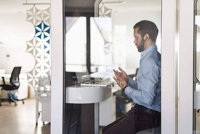 Businessman talking on video call over laptop while sitting in telephone booth at office — Stock Photo