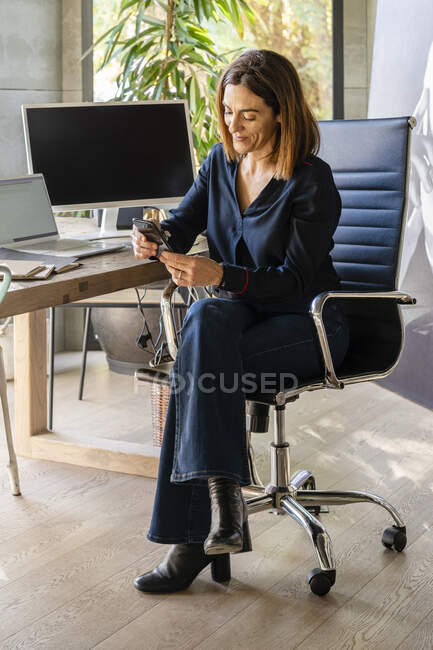 Imprenditrice sorridente che utilizza lo smart phone mentre è seduta sulla sedia in ufficio — Foto stock