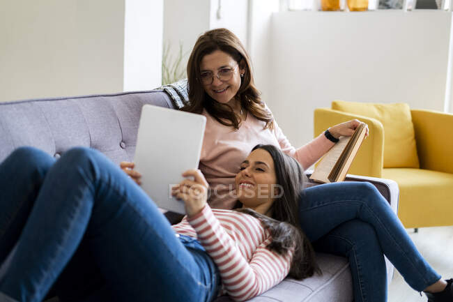 Smiling daughter showing digital tablet while lying on lap of mother with book sitting on sofa at home — Stock Photo