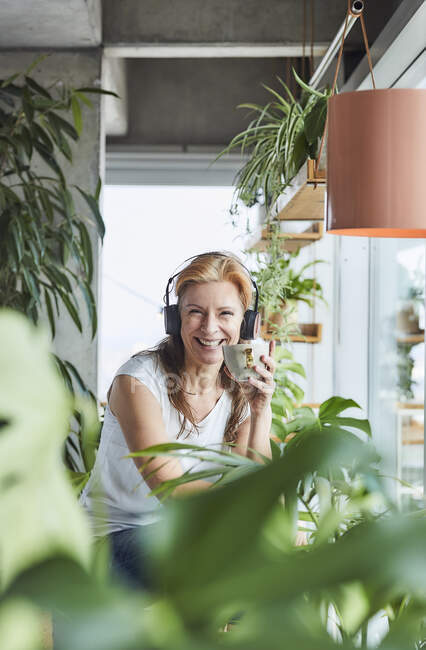 Femme heureuse tenant une tasse de café tout en écoutant de la musique à travers des écouteurs tout en étant assis dans l'appartement loft — Photo de stock