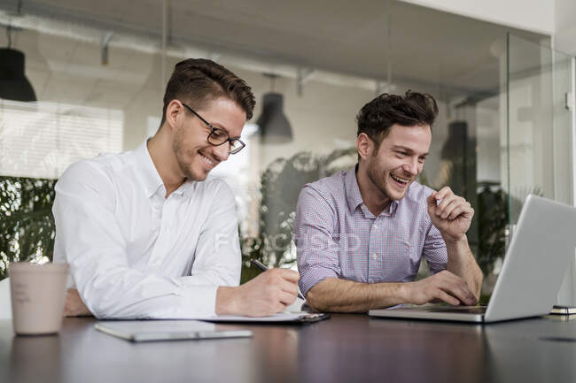Smiling businessman working laptop while male colleague writing on clipboard at office — Stock Photo