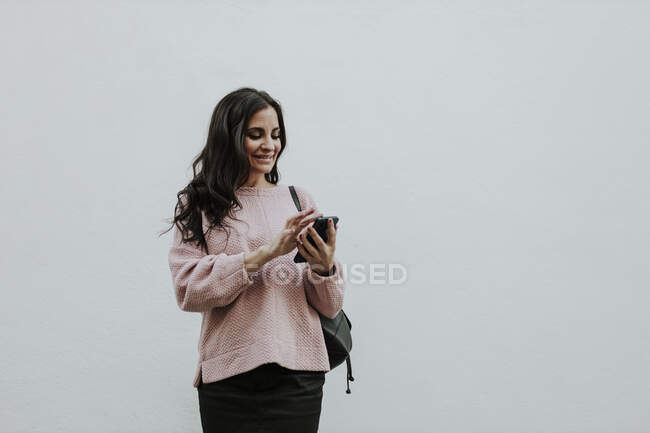 Улыбающаяся женщина с помощью смартфона, стоя напротив белой стены — стоковое фото