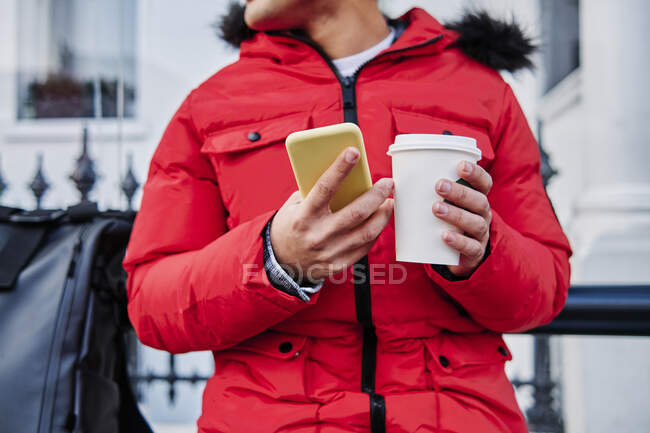 Hombre joven sosteniendo el teléfono móvil y taza de café desechable - foto de stock