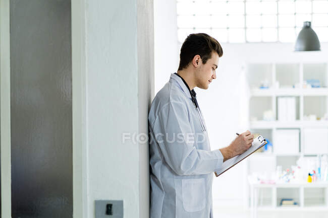 Médico do sexo masculino anotações de escrita profissional enquanto em pé contra a parede no hospital — Fotografia de Stock