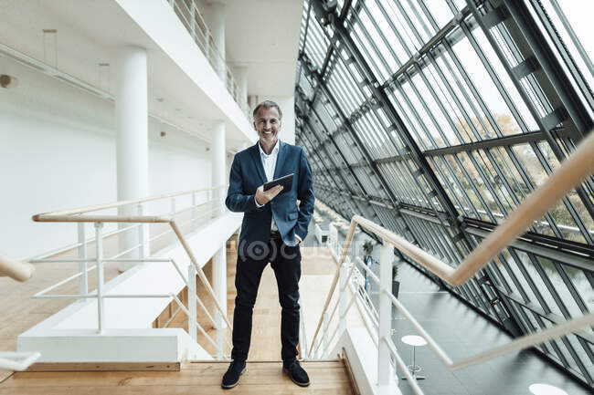 Усміхнений бізнесмен з рукою в кишені тримає цифрову табличку, стоячи в коридорі. — стокове фото