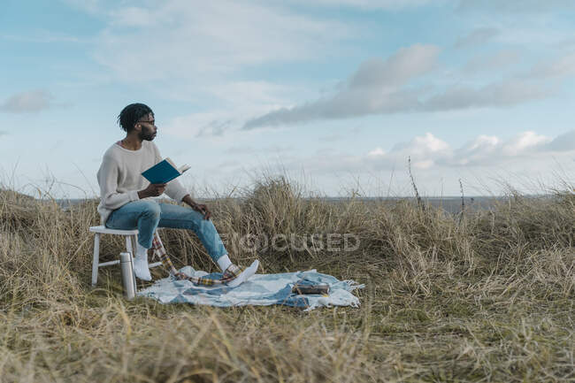 Молодой человек с книгой смотрит в сторону, сидя на стуле против облачного неба — стоковое фото