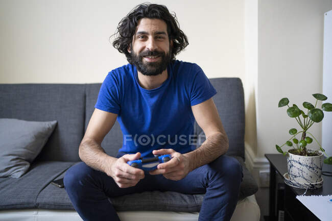Hombre sonriente con controlador de juego sentado en el sofá en casa - foto de stock