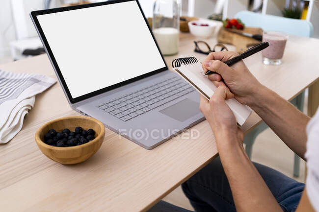 Man sitting with laptop at table while writing on note pad in kitchen at home — Stock Photo