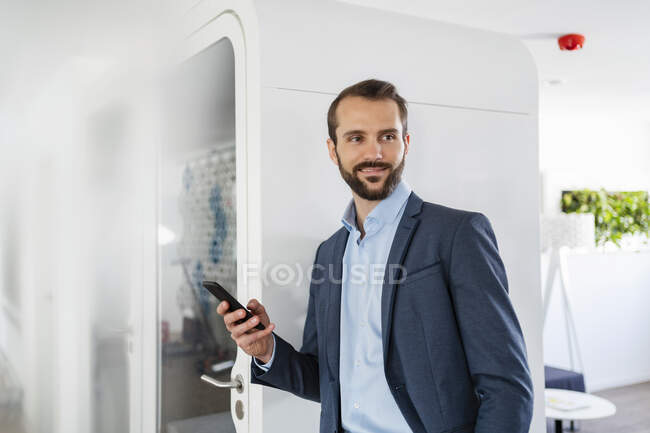 Businessman with mobile phone smiling while standing by telephone booth in office — Stock Photo