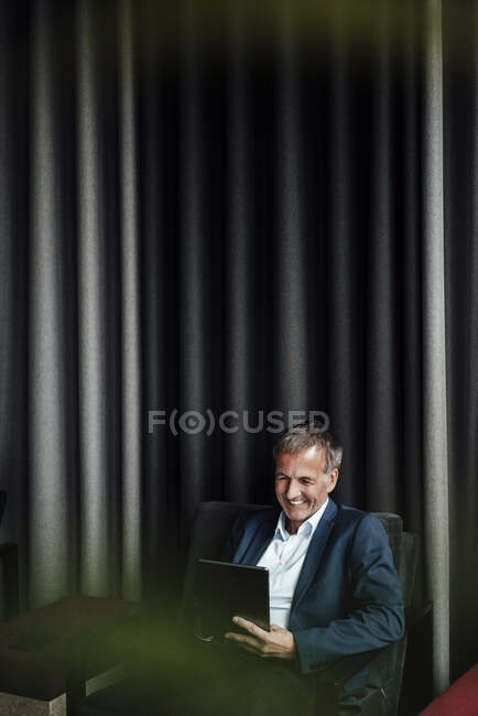 Щасливий старший бізнесмен користується цифровою табличкою, сидячи на стільці проти завіси в офісній кафетерії. — стокове фото