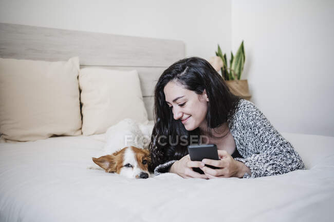 Donna sorridente con cellulare sdraiata da Jack Russell Terrier sul letto di casa — Foto stock