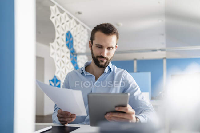 Мужчина-профессионал с бумагой, используя цифровой планшет, сидя в офисе — стоковое фото