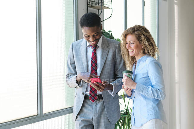Male entrepreneur using smart phone while standing by smiling female colleague with coffee cup — Stock Photo