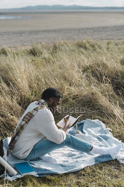 Young man writing in diary while sitting on blanket amidst dried plant at beach — Stock Photo
