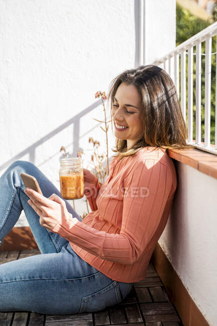 Smiling woman having smoothie while using mobile phone sitting at rooftop — Stock Photo