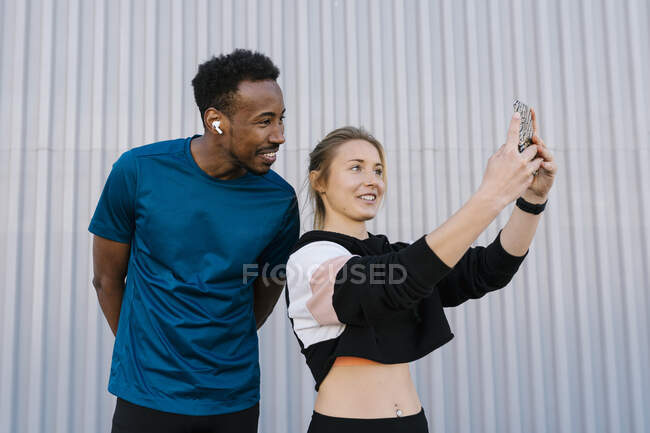 Atleta feminina com homem tirando selfie através do telefone celular enquanto está de pé contra a parede — Fotografia de Stock