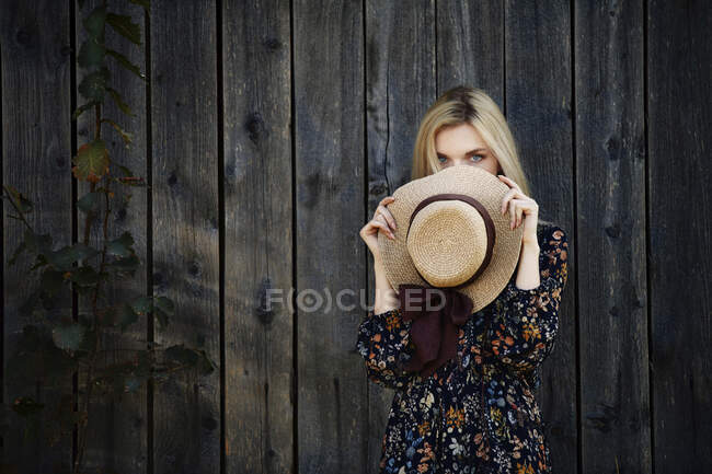 Світла жінка з блакитними очима тримає солом'яний капелюх перед лицем до старої дерев'яної стіни. — стокове фото