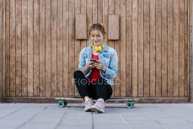 Mujer sonriente con auriculares usando un teléfono inteligente mientras está sentado en el longboard contra la pared de madera - foto de stock