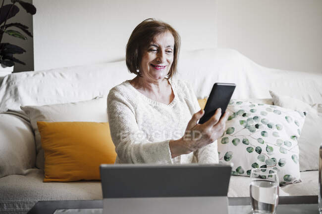 Усміхнена жінка використовує мобільний телефон, сидячи на дивані вдома — стокове фото
