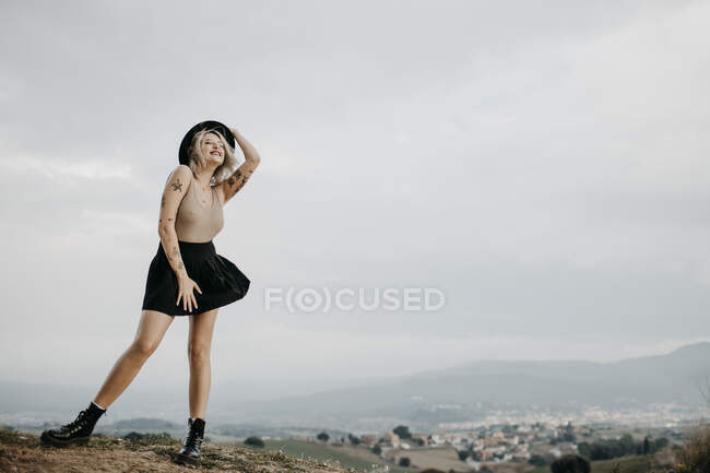 Щаслива молода жінка в капелюсі стоїть на вершині гори проти неба. — стокове фото