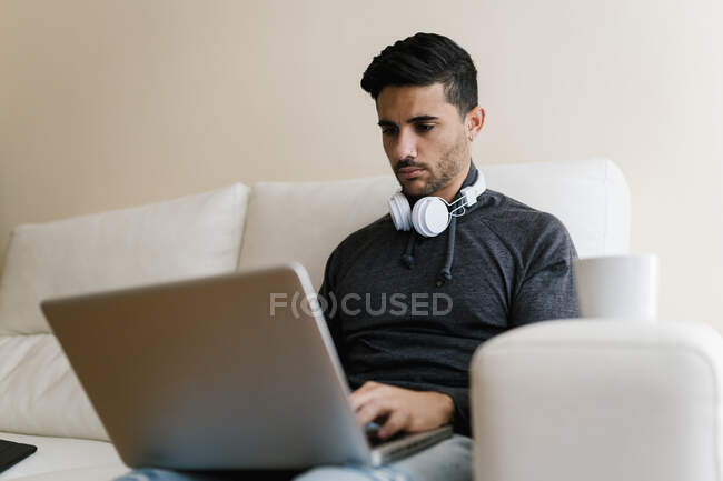 Hombre joven con auriculares usando portátil mientras está sentado en el sofá en casa - foto de stock