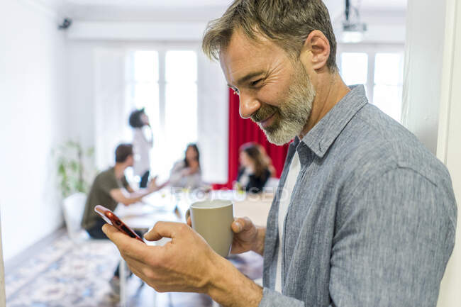 Empreendedor masculino sorridente usando telefone inteligente durante coffee break no escritório — Fotografia de Stock