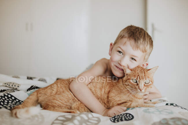 Ragazzo sorridente appoggiato sul gatto rossiccio in camera da letto a casa — Foto stock