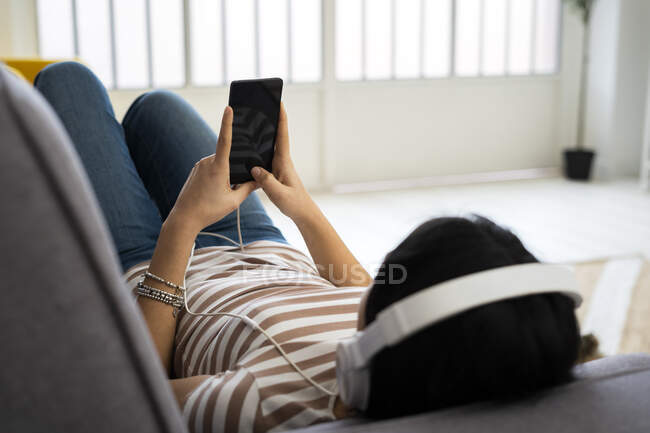 Розслаблена молода жінка слухає музику через бездротові навушники, лежачи на дивані вдома — стокове фото
