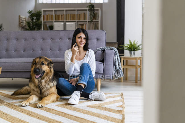 Jovem mulher falando no telefone celular enquanto sentado cão de estimação contra sofá na sala de estar — Fotografia de Stock