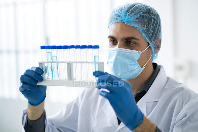 Ученый мужского пола носит защитную маску для лица во время исследования химических веществ в пробирке в лаборатории — стоковое фото