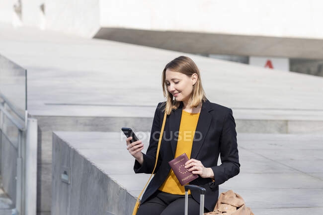 Entrepreneur with passport using mobile phone while sitting on retaining wall — Stock Photo